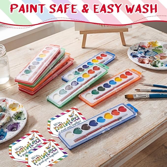 Heart-shaped watercolor paint sets in vibrant colors are arranged on a wooden table, featuring eight hues in each container. Additional paint palettes and brushes are visible, along with cheerful promotional cards highlighting the product.