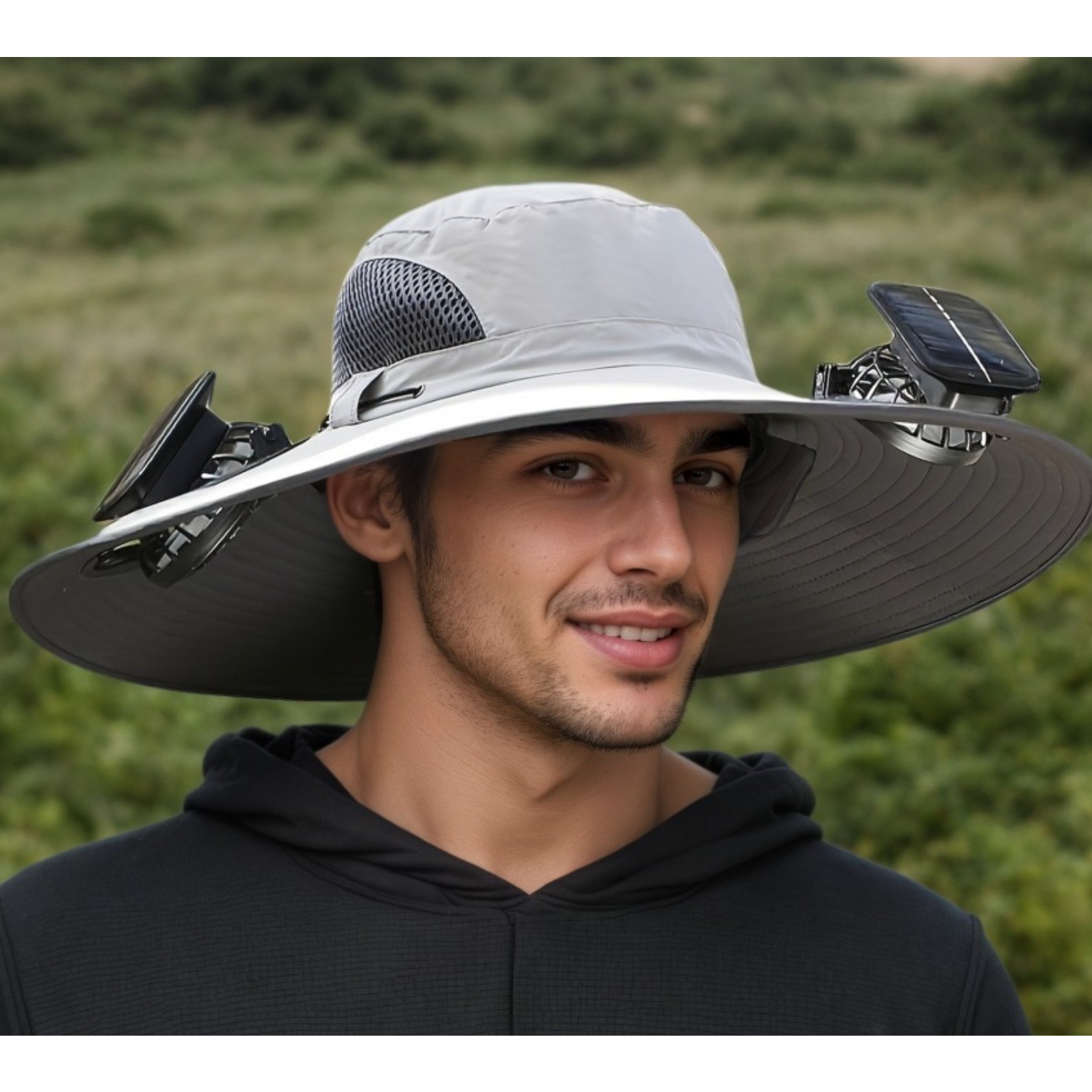 The hat features a wide brim and is primarily grey with solar panels attached on each side, providing ventilation and sun protection. The model, wearing a black hoodie, is standing outdoors in a green landscape.