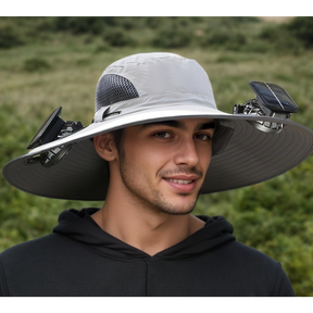 The hat features a wide brim and is primarily grey with solar panels attached on each side, providing ventilation and sun protection. The model, wearing a black hoodie, is standing outdoors in a green landscape.