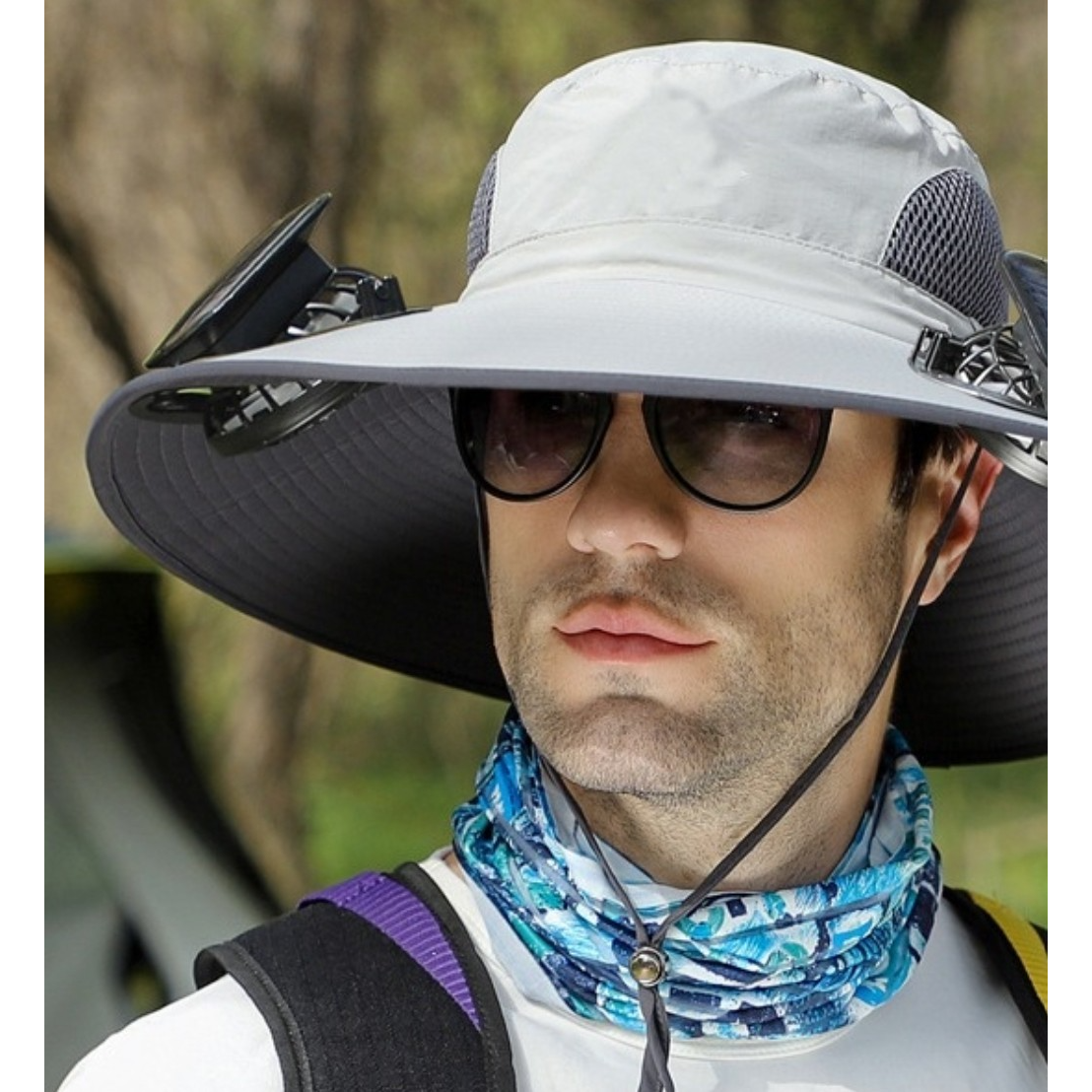 A man wears a light-colored wide-brimmed hat with built-in fans on the sides, sunglasses, and a colorful neck gaiter. The setting appears outdoors with blurred trees in the background.
