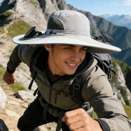 A person wearing a light gray wide-brimmed hat with solar panels on either side is hiking on a rocky trail, surrounded by mountains and greenery in the background. The hat features mesh panels for ventilation and a relaxed fit, contributing to a stylish outdoor look.