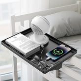 The modern clamp-on floating nightstand features a sleek black tray holding a white mini fan, a book, a pair of glasses, and a smartphone charging with a visible charging indicator. The shelf is securely attached to a bed frame next to a cozy pillow and bedding.