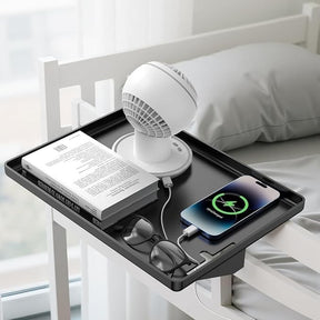 The modern clamp-on floating nightstand features a sleek black tray holding a white mini fan, a book, a pair of glasses, and a smartphone charging with a visible charging indicator. The shelf is securely attached to a bed frame next to a cozy pillow and bedding.