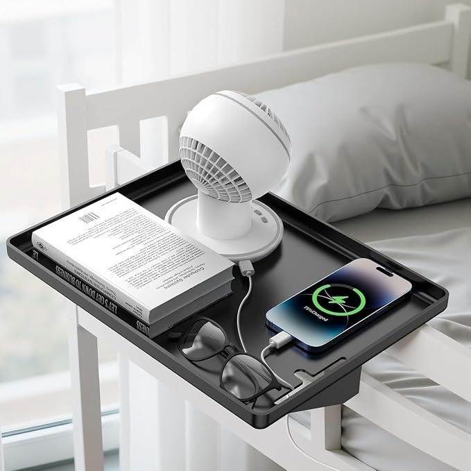 The modern clamp-on floating nightstand features a sleek black tray holding a white mini fan, a book, a pair of glasses, and a smartphone charging with a visible charging indicator. The shelf is securely attached to a bed frame next to a cozy pillow and bedding.