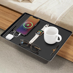 A clamp-on bedside shelf displaying a smartphone with a charging indicator, an Apple Watch, a white cup, AirPods, and a pair of glasses, all resting on a sleek black surface.