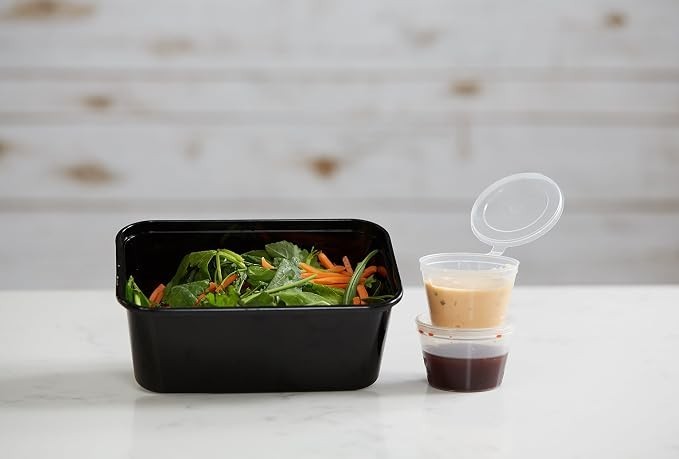 A black rectangular container filled with mixed greens and shredded carrots sits next to two small clear condiment containers, one with a creamy dressing and the other with a dark sauce, both featuring hinged lids.