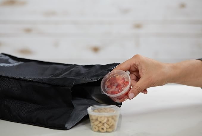 Hand placing a clear container with diced vegetables into a black storage bag, alongside another container filled with cereal.