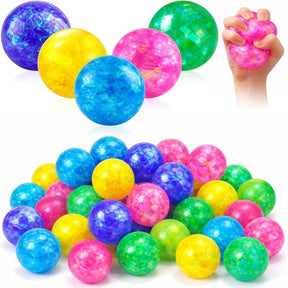 A variety of 20 vibrant glitter stress balls in colors including blue, pink, yellow, green, and purple, displayed in a pile with one being squeezed by a hand. The balls have a glossy, translucent finish showcasing their glittery interior.