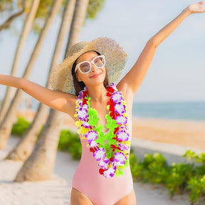 A woman in a pink swimsuit wearing a colorful lei necklace stands in a beach setting, raising her arms joyfully. She is also wearing a wide-brimmed straw hat and oversized white sunglasses, with palm trees and a sandy beach in the background.