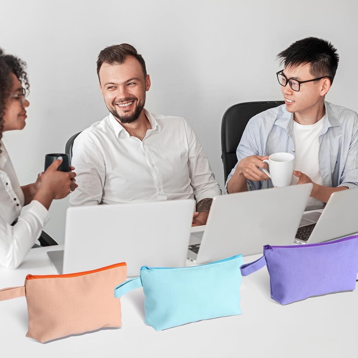 Three zippered pouches in assorted colors — peach with orange zipper, light blue, and purple — are arranged on a table with laptops, while three people engage in conversation in the background.