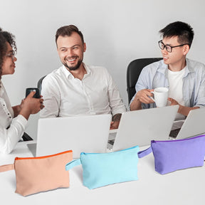 Three zippered pouches in assorted colors — peach with orange zipper, light blue, and purple — are arranged on a table with laptops, while three people engage in conversation in the background.
