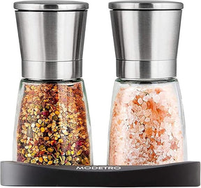 Stylish salt and pepper shakers set featuring a sleek stainless steel top and clear glass bodies, one filled with coarse pink salt and the other with a colorful spice blend, resting on a black stand.