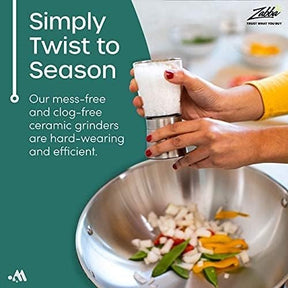 A person is using a salt shaker over a metal bowl filled with colorful sliced vegetables, demonstrating the product's functioning. The background features brushed metal and out-of-focus greenery, enhancing the kitchen setting.