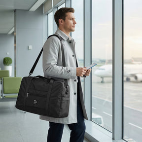 A man stands at an airport window holding a boarding pass, wearing a light gray coat with the black foldable weekender tote bag slung over his shoulder. The bag features a front zip pocket and is set against a backdrop of planes at the terminal.