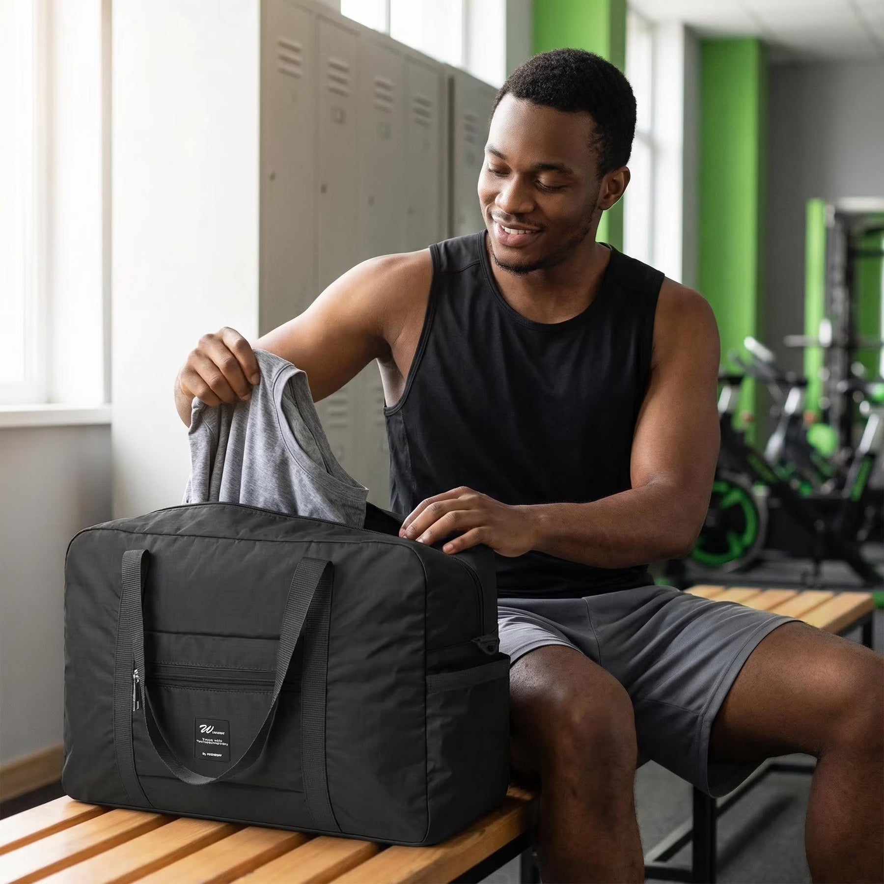A man in a black tank top is sitting on a bench in a gym, placing a gray shirt into a black foldable tote bag with dual handles and a front pocket. The setting features lockers and exercise equipment in the background.