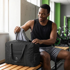 A man in a black tank top is sitting on a bench in a gym, placing a gray shirt into a black foldable tote bag with dual handles and a front pocket. The setting features lockers and exercise equipment in the background.