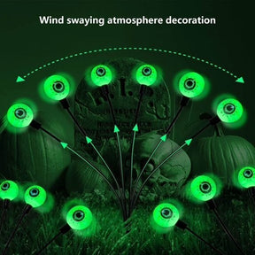 Eight green LED eyeball lights are arranged in a spread-out pattern on black wires, glowing against a dark background featuring a tombstone, skull, and pumpkins, creating a spooky Halloween atmosphere.