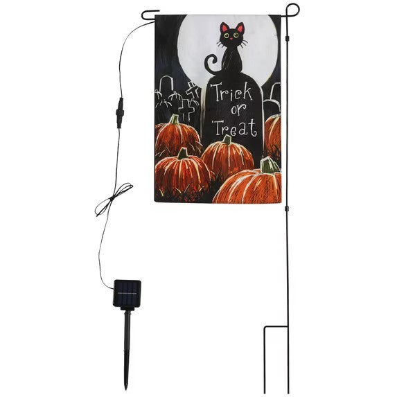 Colorful garden flag featuring a black cat in front of a full moon, with pumpkins and the text "Trick or Treat" displayed prominently. The flag is attached to a black metal stake with a solar panel connected below.