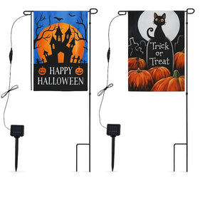 Two Halloween-themed solar garden flags are displayed; the left features a haunted house under a full moon with "Happy Halloween" text, while the right shows a black cat beside a "Trick or Treat" tombstone and pumpkins. Each flag is mounted on a black metal stand with a solar panel at the base for illumination.