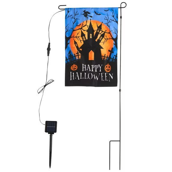 The garden flag features a Halloween theme with a black background, an orange moon, a spooky silhouette of a haunted house, and "HAPPY HALLOWEEN" text in white, displayed on a metal stand alongside a solar panel for charging.