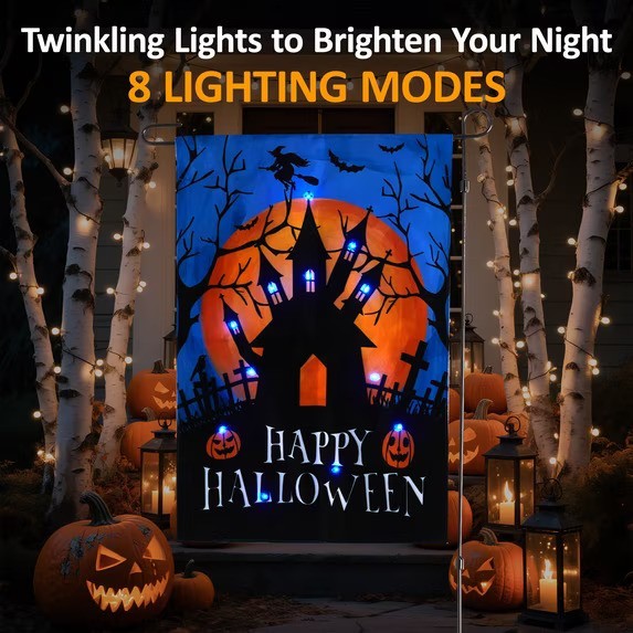 Halloween-themed garden flag featuring a spooky haunted house design with a large orange moon and a flying witch. The flag displays the text "HAPPY HALLOWEEN" and includes blue LED lights, surrounded by decorative pumpkins and lanterns in a festive outdoor setting.