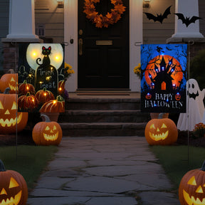 Two illuminated garden flags display Halloween themes: one featuring a black cat with the text "Trick or Treat" and the other depicting a haunted house with "Happy Halloween" against a glowing orange moon. Surrounding the flags are carved pumpkins, adding to the festive atmosphere.