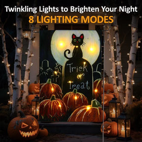 A colorful Halloween-themed garden flag featuring a black cat in front of a gravestone with "Trick or Treat" written on it, surrounded by glowing pumpkins. The flag is illuminated with twinkling lights, set against a backdrop of trees and lanterns that enhance the festive atmosphere.