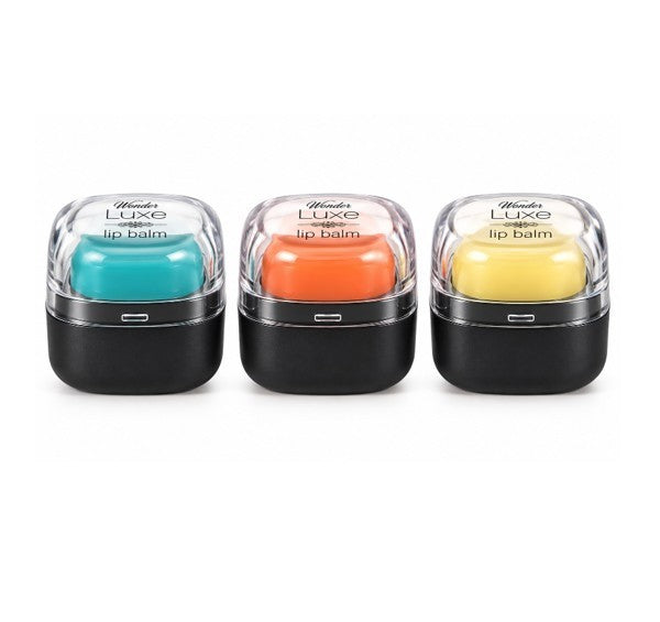 Three round lip balm containers with clear lids showcasing teal, orange, and yellow balms, each sitting in sleek black bases.