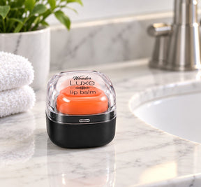 The lip balm is shown in a clear, dome-shaped container with a black base, revealing a vibrant orange balm inside. The setting features a marble countertop with a soft white towel and a plant in the background.