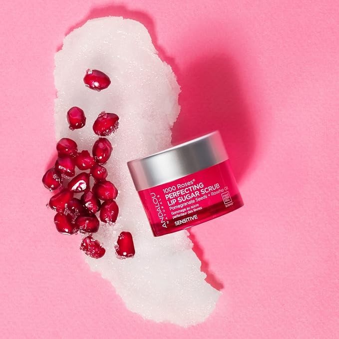The container of the 1000 Roses Perfecting Lip Sugar Scrub is positioned on a bed of white sugar, surrounded by shiny pomegranate seeds, against a bright pink background. The jar has a silver lid with a vibrant pink label showcasing the product details.