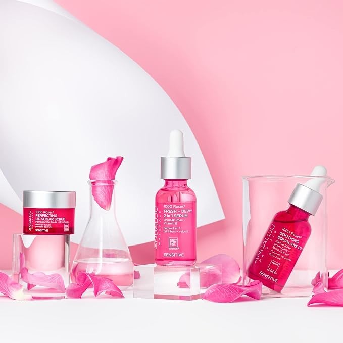 Arrangement of Andalou Naturals products on a pink background, featuring the 1000 Roses Perfecting Lip Sugar Scrub in a small jar on the left, a pink dropper bottle of Fresh + Dew 2-in-1 Serum in the center, and a Soothing Squalane Oil bottle on the right, surrounded by rose petals.