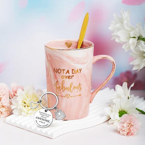 A pink marbled mug with gold lettering reading "NOT A DAY over fabulous" is accompanied by a gold spoon and a silver keychain featuring a heart and messages about being awesome, arranged on a textured white dish. Surrounding florals add a decorative touch.