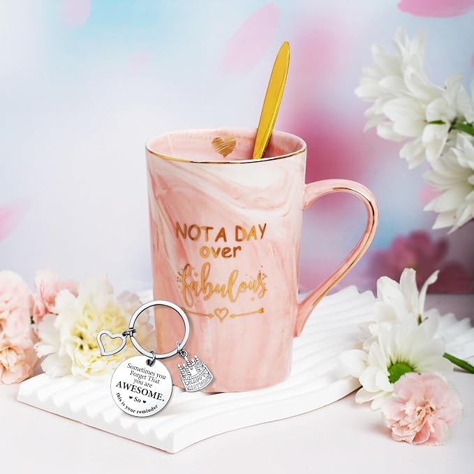 A pink marbled mug with gold lettering reading "NOT A DAY over fabulous" is accompanied by a gold spoon and a silver keychain featuring a heart and messages about being awesome, arranged on a textured white dish. Surrounding florals add a decorative touch.
