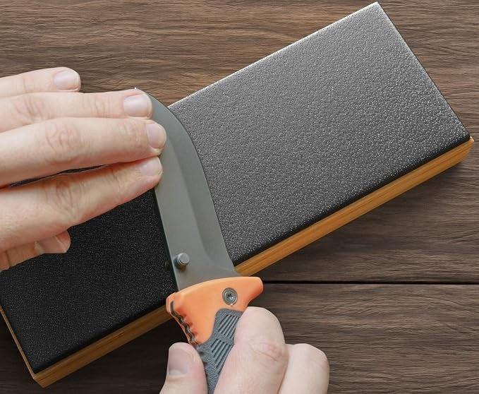 A hand holding a folding knife gently sharpens its blade against the textured black surface of the Sharp Pebble Gladius Leather Strop Sharpener, which features a wooden base. The setting has a natural wood grain background.