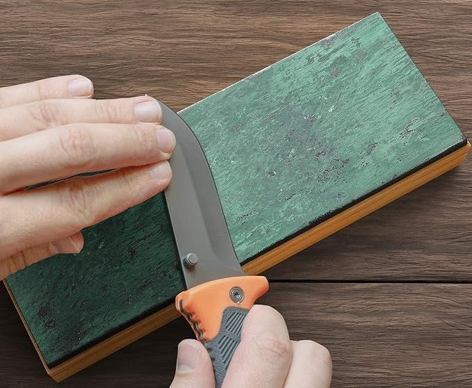A hand is shown holding a curved knife against a double-sided leather strop with a green surface, placed on a wooden table. The strop features a smooth edge and a wooden base, illustrating its use for sharpening.