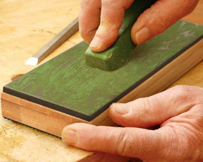 A hand holds a green stropping compound against a double-sided leather strop sharpener resting on a wooden surface. The strop features a textured green surface and a smooth black side, with visible wood grain on the base.