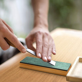 A person is using a sharp knife on a green leather strop, which is resting on a wooden table. The bamboo base of the strop is visible, highlighting its texture and color.
