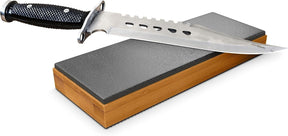 The leather strop sharpener rests on a bamboo base with a rectangular grey stropping surface, and a sleek knife with a black textured handle is positioned on top, angled slightly to the side.