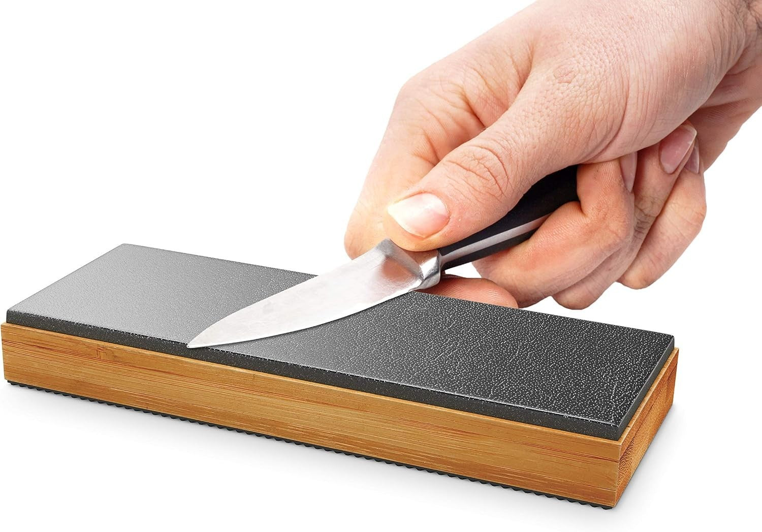 A hand is holding a knife against the double-sided black leather surface of the Sharp Pebble Gladius Leather Strop Sharpener, which features a durable bamboo base. The sharpener is oriented horizontally and shows a textured surface for effective sharpening.