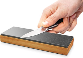 A hand is holding a knife against the double-sided black leather surface of the Sharp Pebble Gladius Leather Strop Sharpener, which features a durable bamboo base. The sharpener is oriented horizontally and shows a textured surface for effective sharpening.