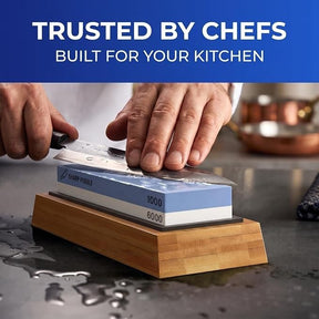 A person's hand holds a knife against a blue Sharp Pebble whetstone labeled with 1000 and 6000 grit, positioned on a bamboo base, with water droplets on the stone and countertop. The background features kitchen utensils and a copper pot.