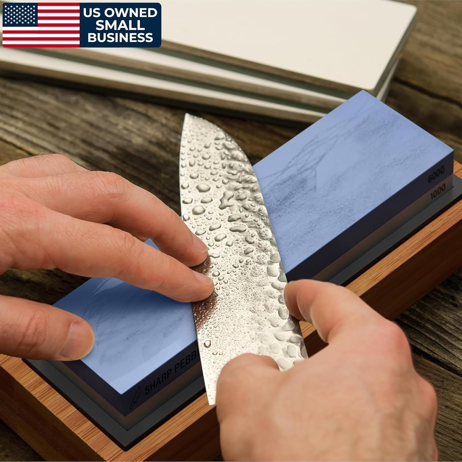 A hand holds a knife with water droplets on its blade, positioned over a dual-sided blue whetstone labeled 1000 and 6000 grit, resting on a wooden base. The setting features a rustic wooden table with additional sharpening stones partially visible in the background.