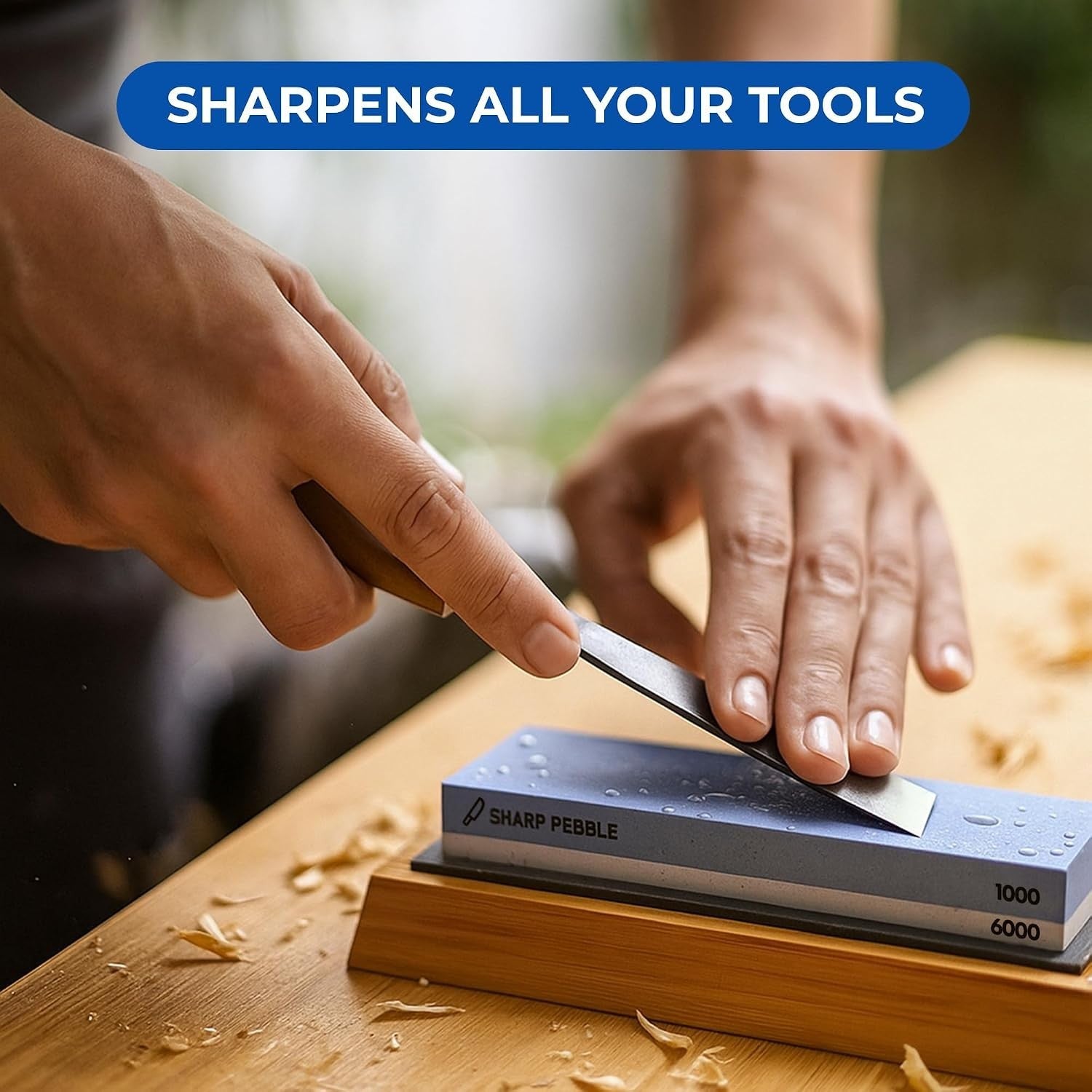 A person is sharpening a knife on a blue Sharp Pebble whetstone, with visible water droplets on the stone and wood shavings scattered on a wooden surface. The whetstone is displaying both 1000 and 6000 grit sides.