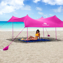 Large pink beach tent with a sleek, modern design, positioned on sand near the ocean. A woman in a yellow bikini relaxes underneath, lounging on a blue blanket while wearing sunglasses.