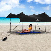 A large black beach tent is set up on the sand, providing sun shade near the ocean. A woman in a yellow bikini relaxes on a blue blanket beneath the canopy.