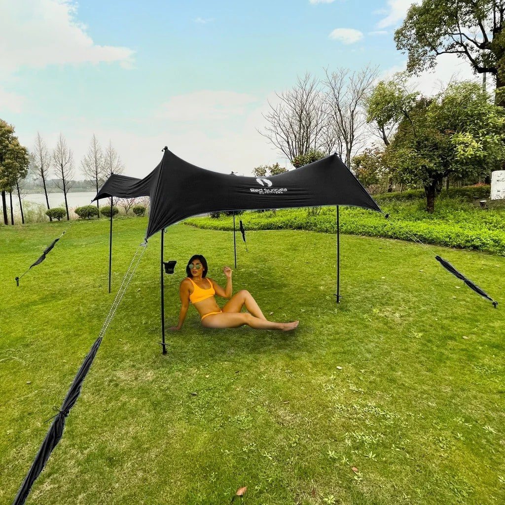 A woman in a yellow bikini relaxes on a blue mat beneath a large bright pink beach tent on a sandy beach, with the ocean and blue sky in the background. The tent features multiple poles and guy lines for stability.