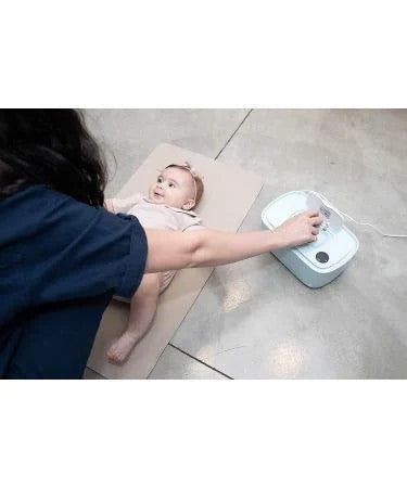 A caregiver is reaching for the Easy Baby Multi-Functional Wipes Heater, which is light blue, while a happy baby lays on a soft surface nearby. The image captures a cozy, nurturing moment between the caregiver and the baby.