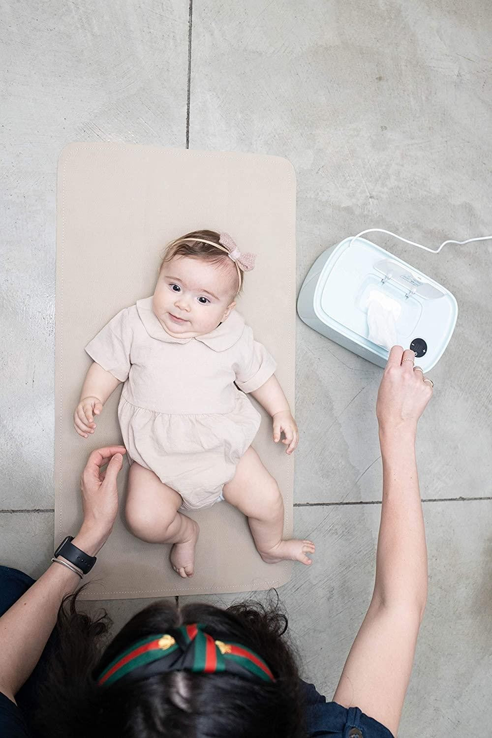 A smiling baby in a light-colored, short-sleeved outfit lies on a beige changing mat, while an adult's hand reaches towards a white wipes heater beside them, with a partially pulled-out wipe visible.