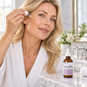 A woman in a white robe applies a drop of Elastalift Collagen Serum to her cheek while smiling, with the bottle of serum visible on a marble countertop beside her. The setting includes a bathroom with natural light and soft greenery in the background.
