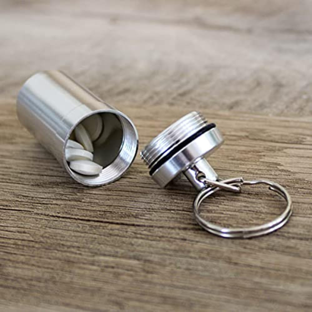 The metal capsule pill box is shown opened, revealing several white tablets inside, with the attached keychain and screw-on lid visible in the foreground. The surface beneath is wooden, providing a natural backdrop.
